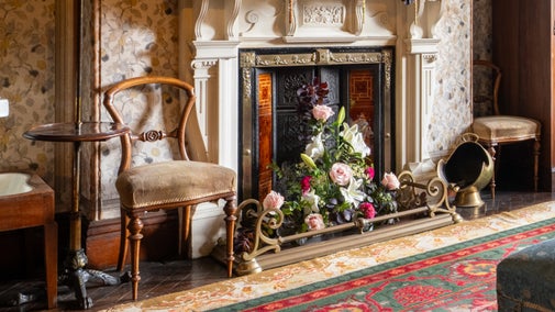 Vintage fireplace dressed with a floral arrangement of roses, lilies and greenery, framed by ornate tiles and surrounded by period furniture in a historic Sunnycroft interior.